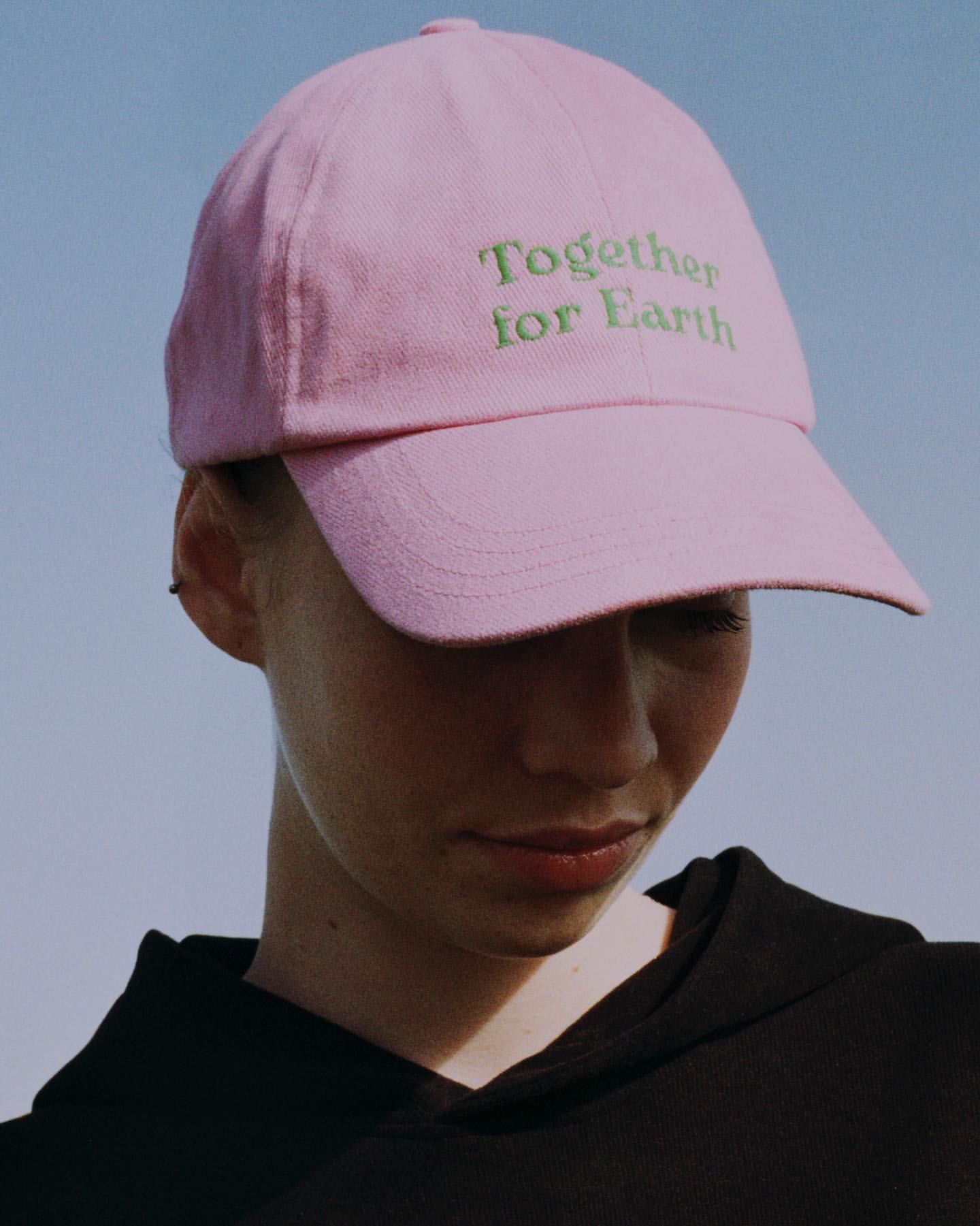 Young woman in a black hoodie wears a pink baseball cap embroidered with 'Together for Earth' against a pale blue sky background.