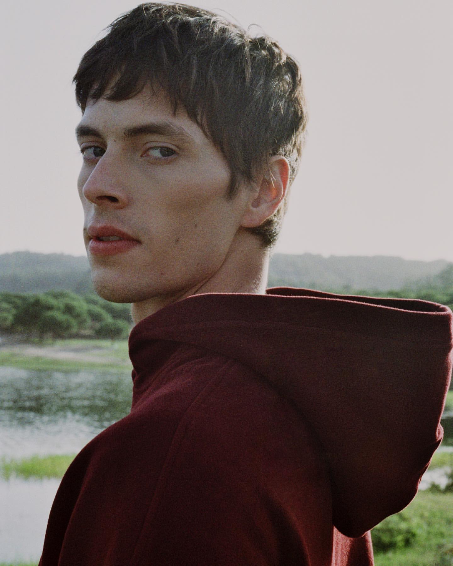 Portrait of a young adult male in a maroon hoodie, looking over his shoulder with a soft, natural outdoor setting in the background.