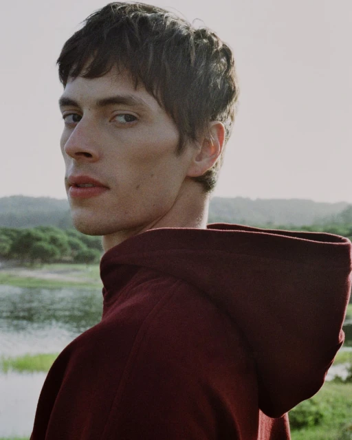 Portrait of a young adult male in a maroon hoodie, looking over his shoulder with a soft, natural outdoor setting in the background.