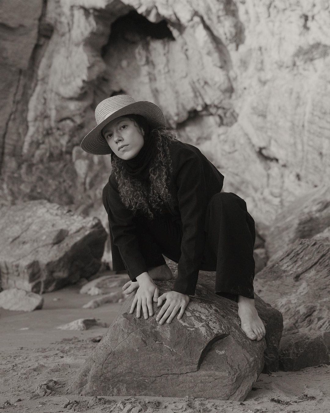 Monochrome portrait of a curly-haired person in a hat, seated on a rock formation near a sandy area with a rocky cliff in the background.