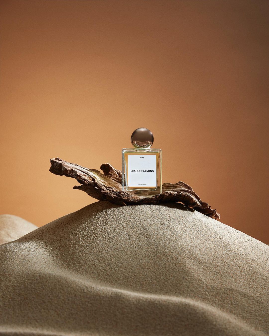 Still life shot of a perfume bottle with a gold cap sitting on a piece of driftwood and sand against a tan background.