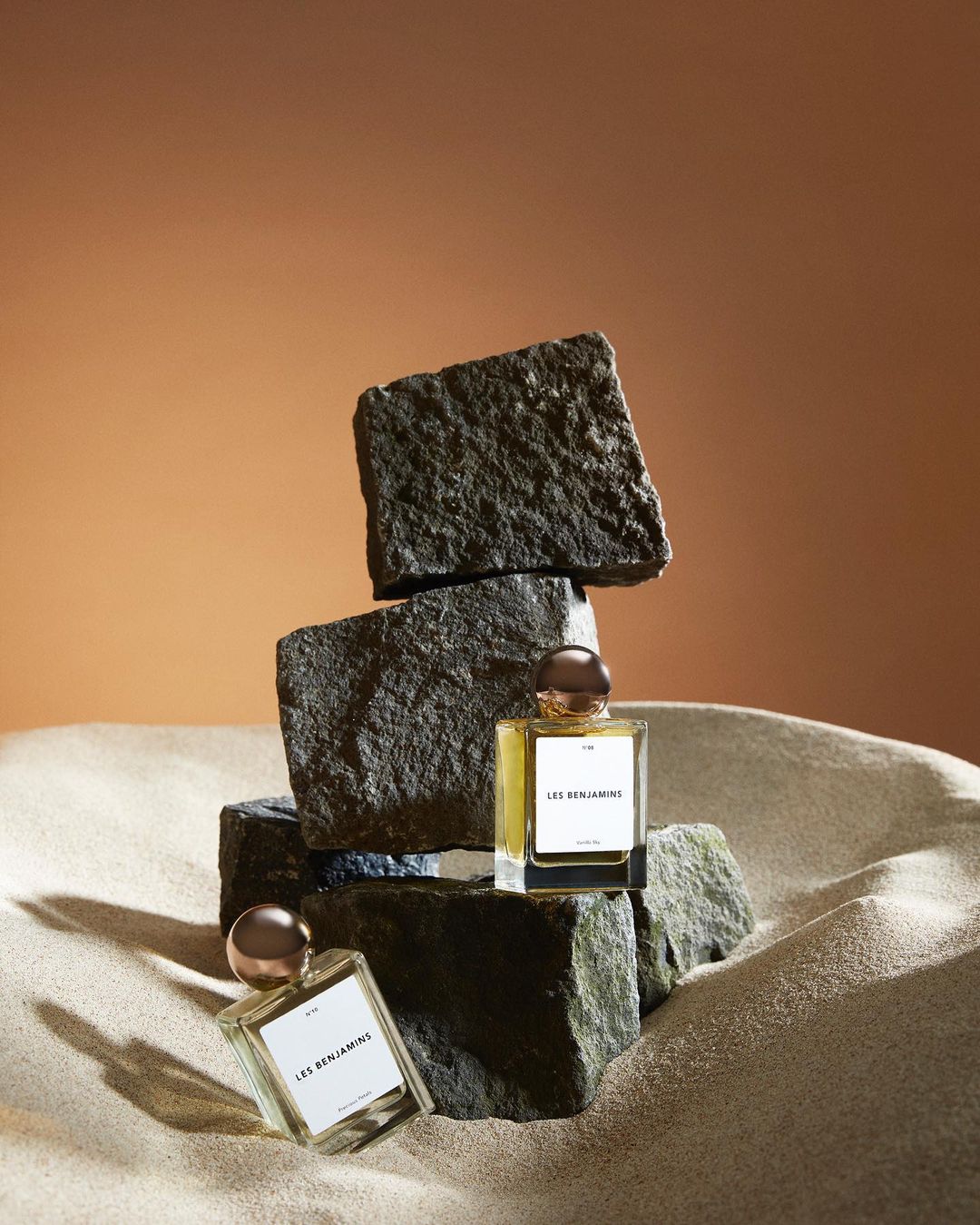 Product shot of two Les Benjamins fragrance bottles displayed on rough stone, against a tan background, emphasizing texture and luxury.