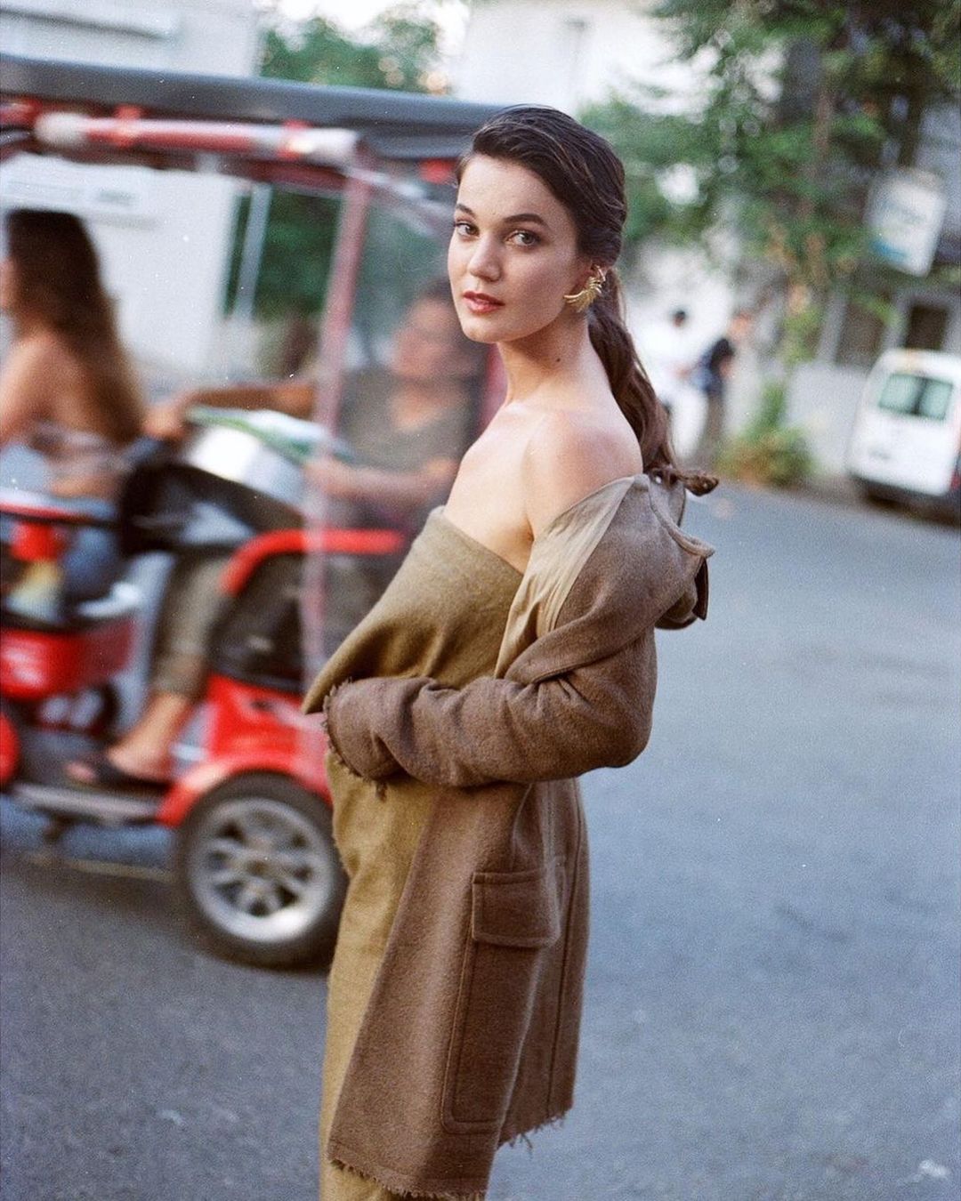 A woman in a brown coat looks over her shoulder on a city street with a red tuk-tuk visible in the background.