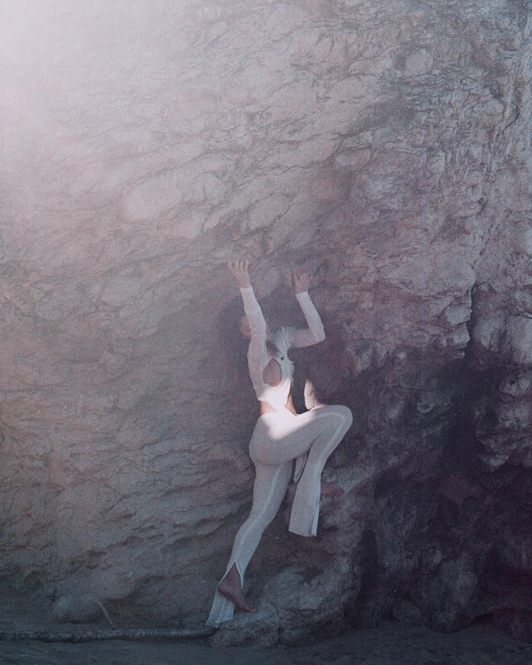 A female figure in white clings to a rough, grey rock face, set against a soft, diffused background, creating a surreal, ethereal image.