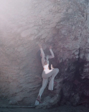A female figure in white clings to a rough, grey rock face, set against a soft, diffused background, creating a surreal, ethereal image.