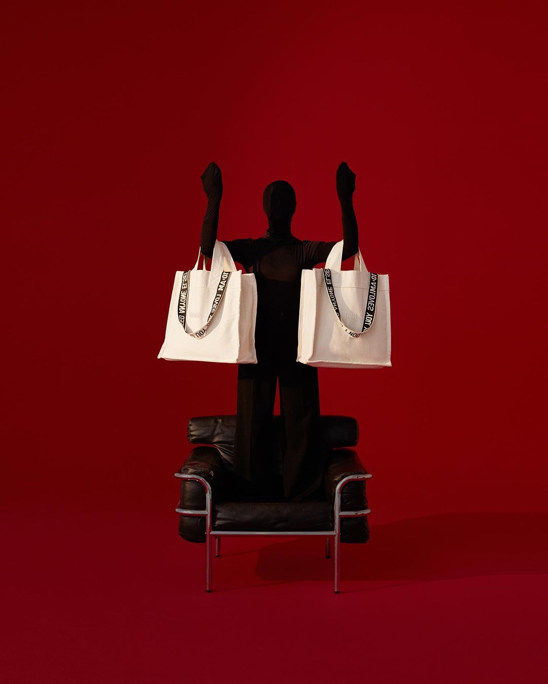 Person in black bodysuit sits in chair holding shopping bags against red background in studio. Minimalist fashion photography.