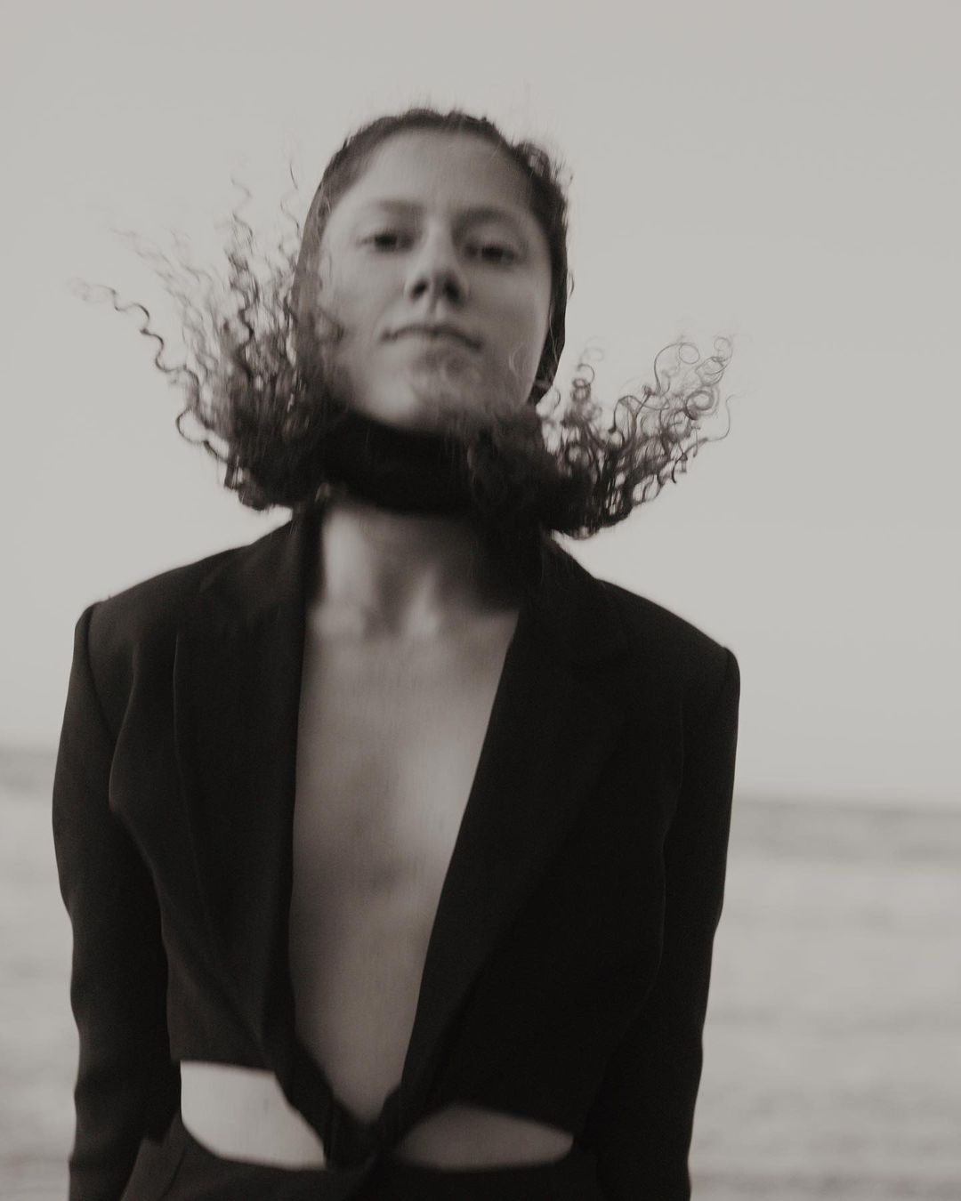 Monochrome portrait of a person with curly hair wearing a dark blazer against an outdoor sky background