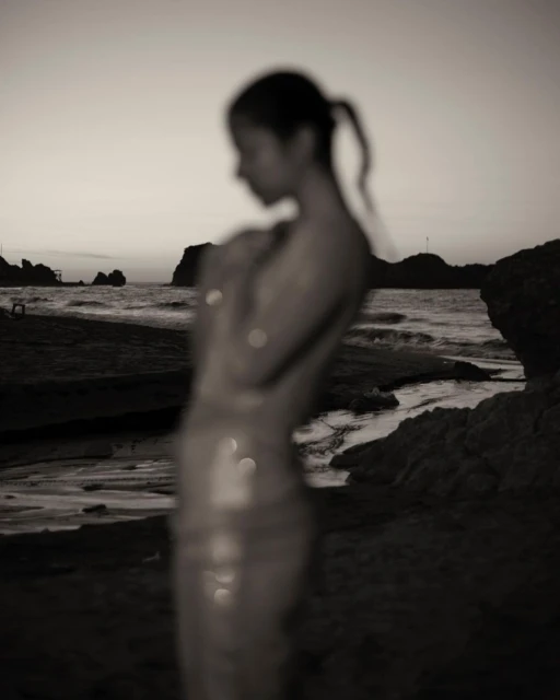 Monochromatic blurred shot of a woman standing at the beach during sunset, with visible rocks and ocean water.