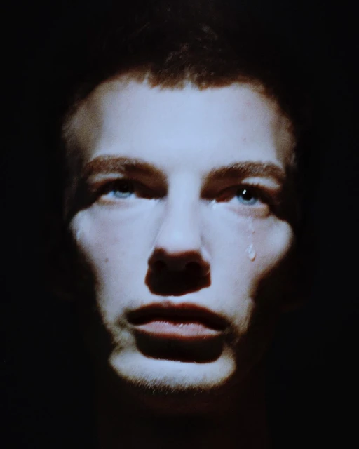 Close-up portrait of a young adult man with a tear running down his face in dramatic, dark lighting, highlighting his blue eyes.