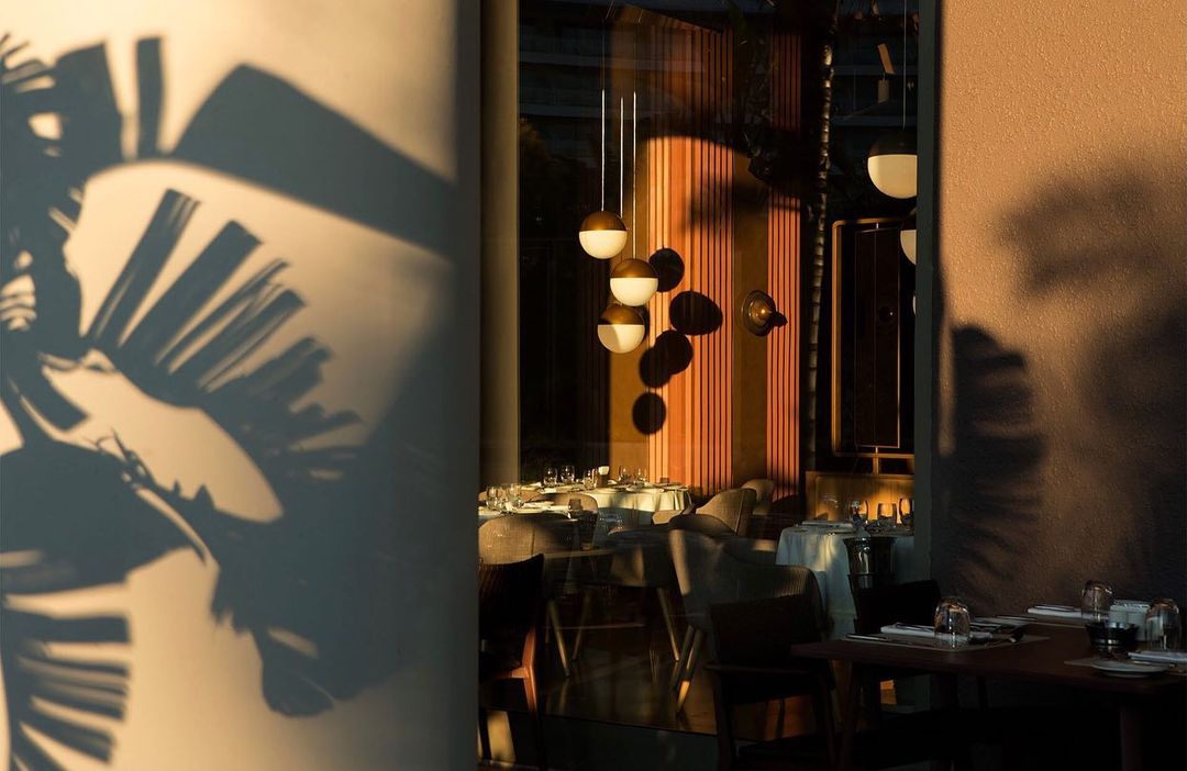Upscale restaurant interior with pendant lights above tables, chairs, place settings, and shadows from foliage, creating a refined atmosphere.