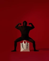 Figure in dark hooded sweatshirt flexing arms over a white tote bag against a solid red background