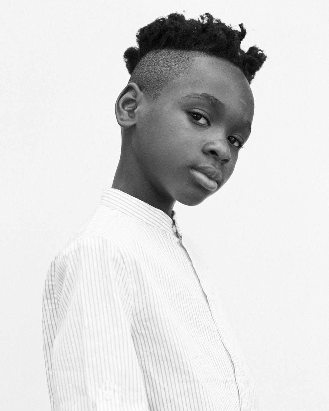Black and white portrait of a young Black person with short hair, wearing a striped white shirt, against a plain white backdrop.