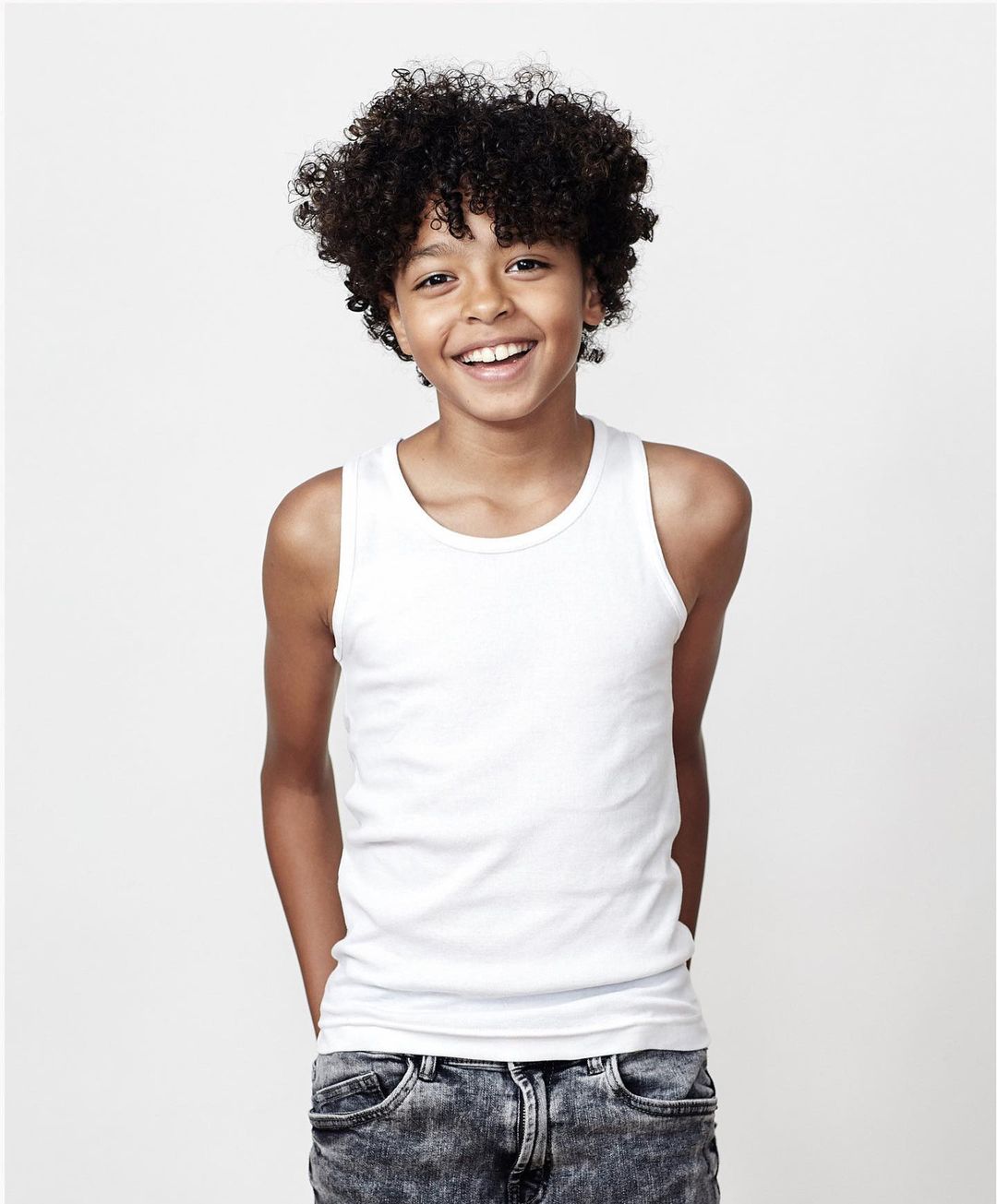A smiling young boy with curly dark hair and fair skin, wearing a white tank top and jeans, poses in front of a neutral background.