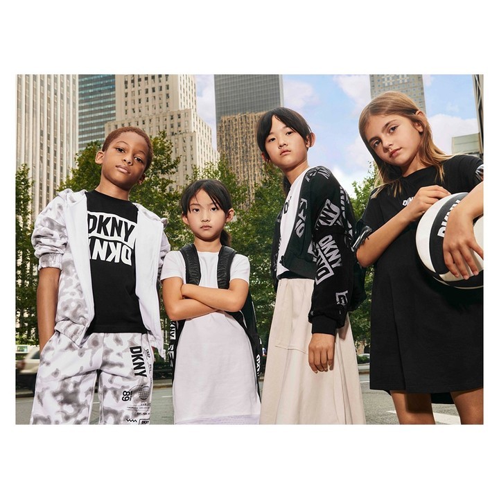 Four children model DKNY clothing on a city street. Two girls and two boys stand close together in casual DKNY branded outfits.
