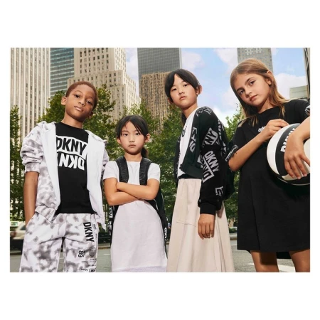 Four children model DKNY clothing on a city street. Two girls and two boys stand close together in casual DKNY branded outfits.