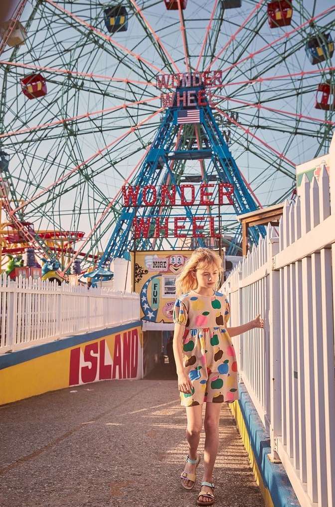 A young girl in a patterned dress walks on a boardwalk near a white fence, with the Wonder Wheel Ferris wheel in the background.
