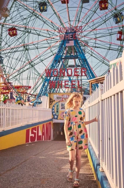 A young girl in a patterned dress walks on a boardwalk near a white fence, with the Wonder Wheel Ferris wheel in the background.