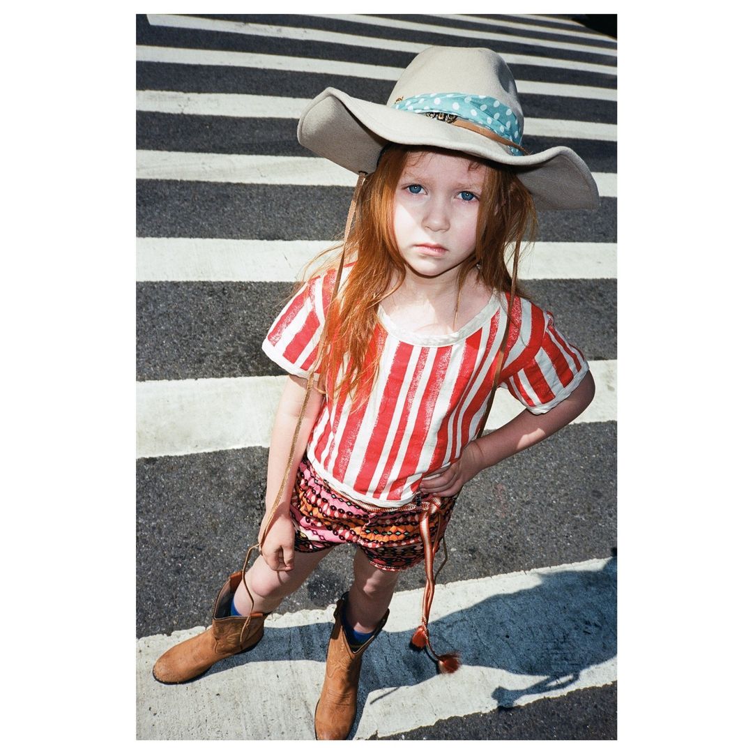 Young girl in cowboy hat and boots stands on crosswalk. She wears a red-and-white striped shirt and patterned skirt in full shot.