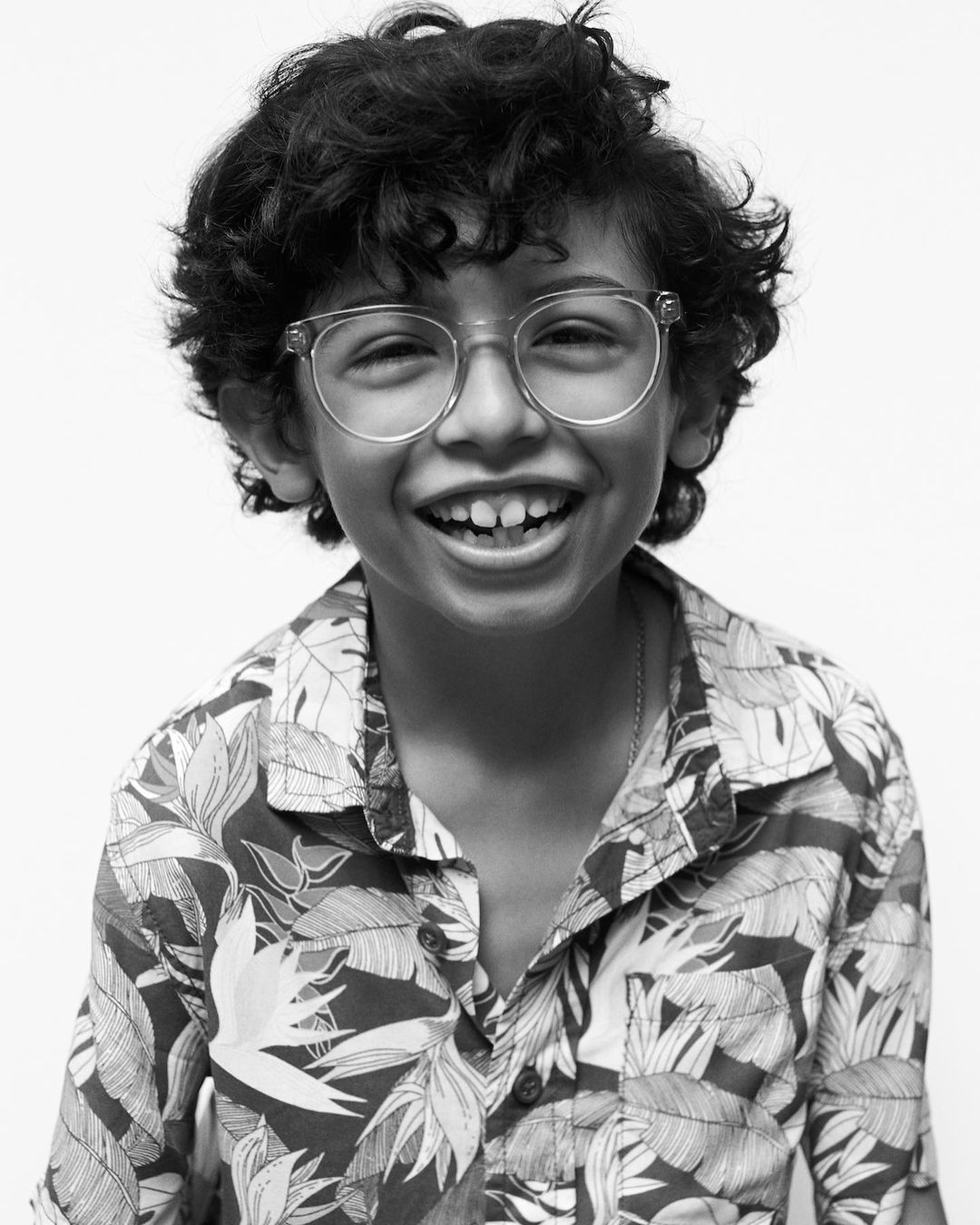 Young boy with curly hair, wearing glasses and a floral shirt, smiles against a plain white backdrop in a close-up black and white portrait.