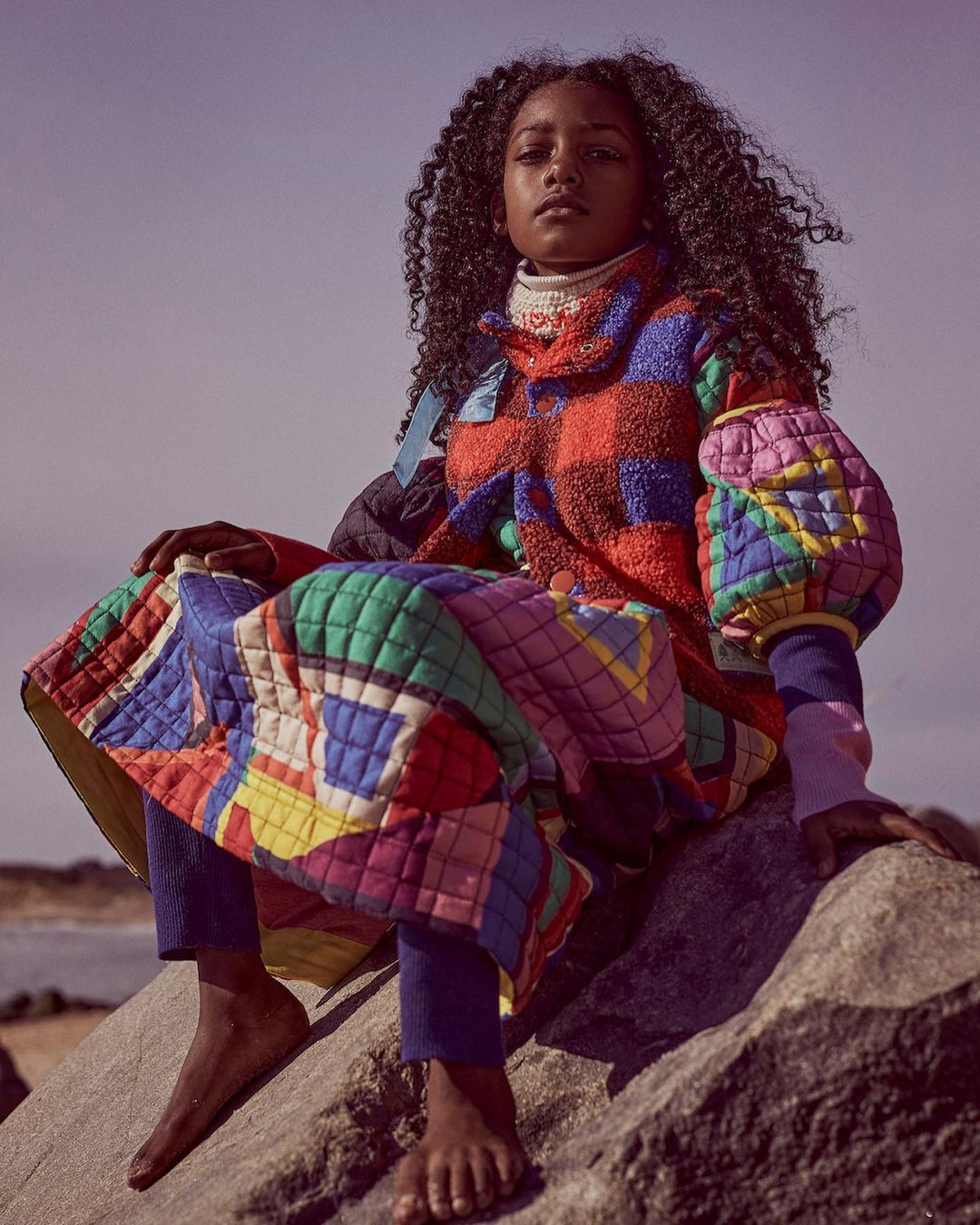 A young, dark-skinned child sits on a rock, wearing a vibrantly colored quilted coat with bare feet, set against a backdrop of a blue sky and beach.