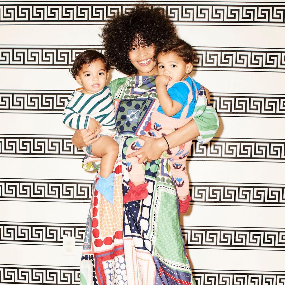 Mother holds two young children in front of a geometric patterned wallpaper wearing a colourful patchwork-style dress and curly hair.