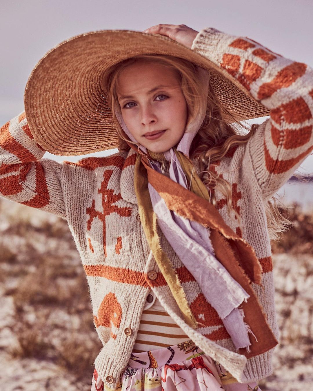 Blonde child model wearing a patterned cardigan, striped shirt, and sun hat poses on a beach with a scarf around her neck.