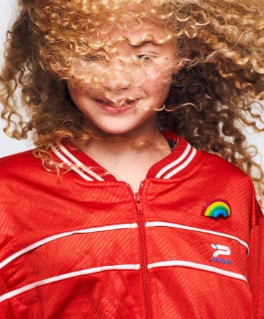Close-up portrait of a child with blonde curly hair covering eyes wearing a red zip-up jacket against a light background