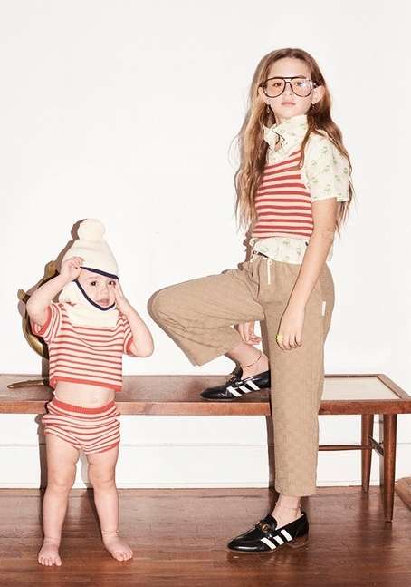 Two children pose on wooden furniture in a brightly lit, indoor scene. One child wears a knit cap, while the other sports oversized glasses.