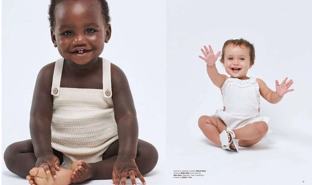 Two adorable babies, one with dark skin and one with light skin, are sitting on a white surface wearing rompers and smiling at the camera.