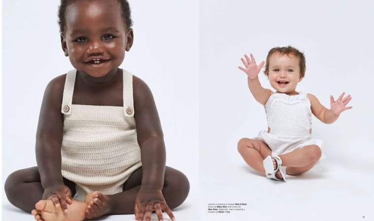 Two adorable babies, one with dark skin and one with light skin, are sitting on a white surface wearing rompers and smiling at the camera.