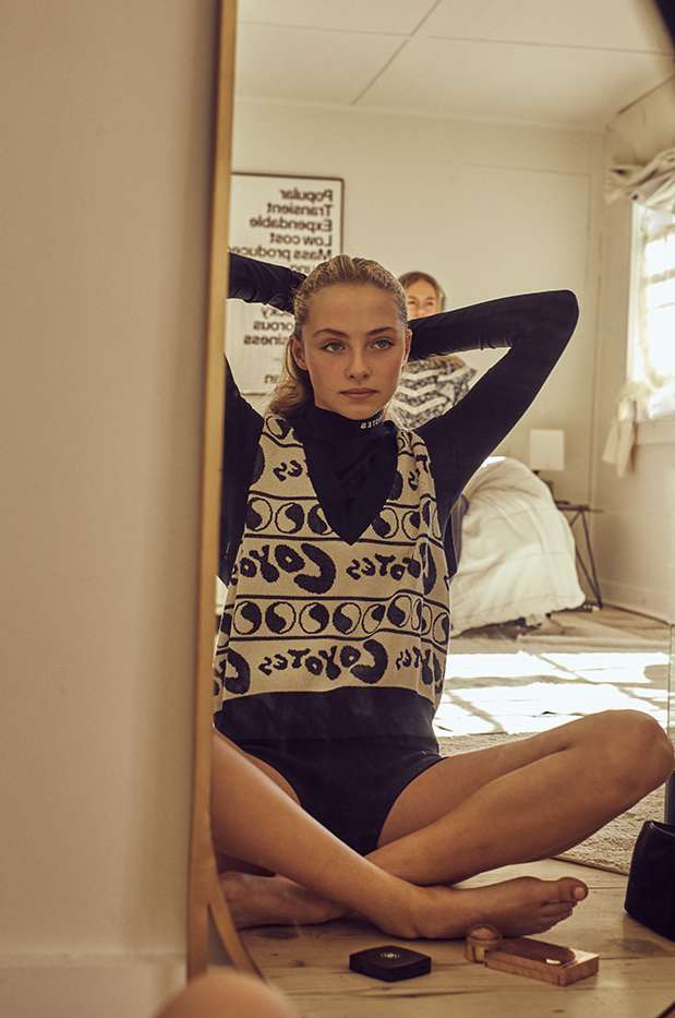 Teen girl sits cross-legged on the floor in front of a mirror, adjusting her hair, with another person and bed visible in the background.