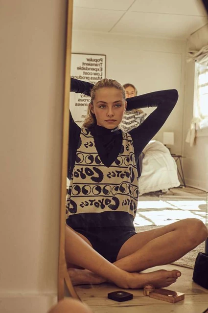 Teen girl sits cross-legged on the floor in front of a mirror, adjusting her hair, with another person and bed visible in the background.