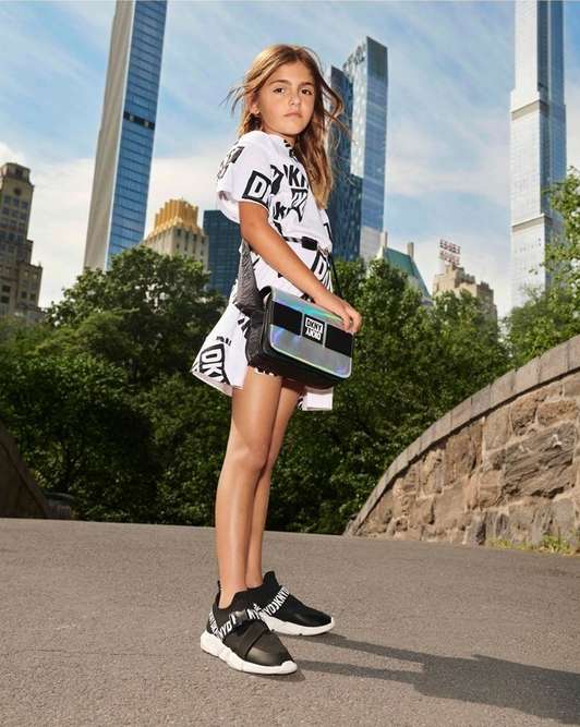 Young girl poses outdoors wearing a white DKNY logo dress, black sneakers, and a shiny silver purse against a city and park background.