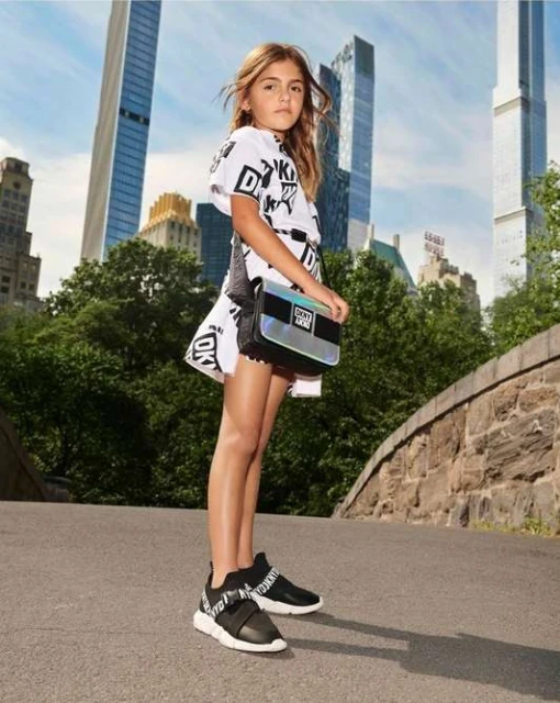 Young girl poses outdoors wearing a white DKNY logo dress, black sneakers, and a shiny silver purse against a city and park background.