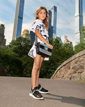 Young girl poses outdoors wearing a white DKNY logo dress, black sneakers, and a shiny silver purse against a city and park background.