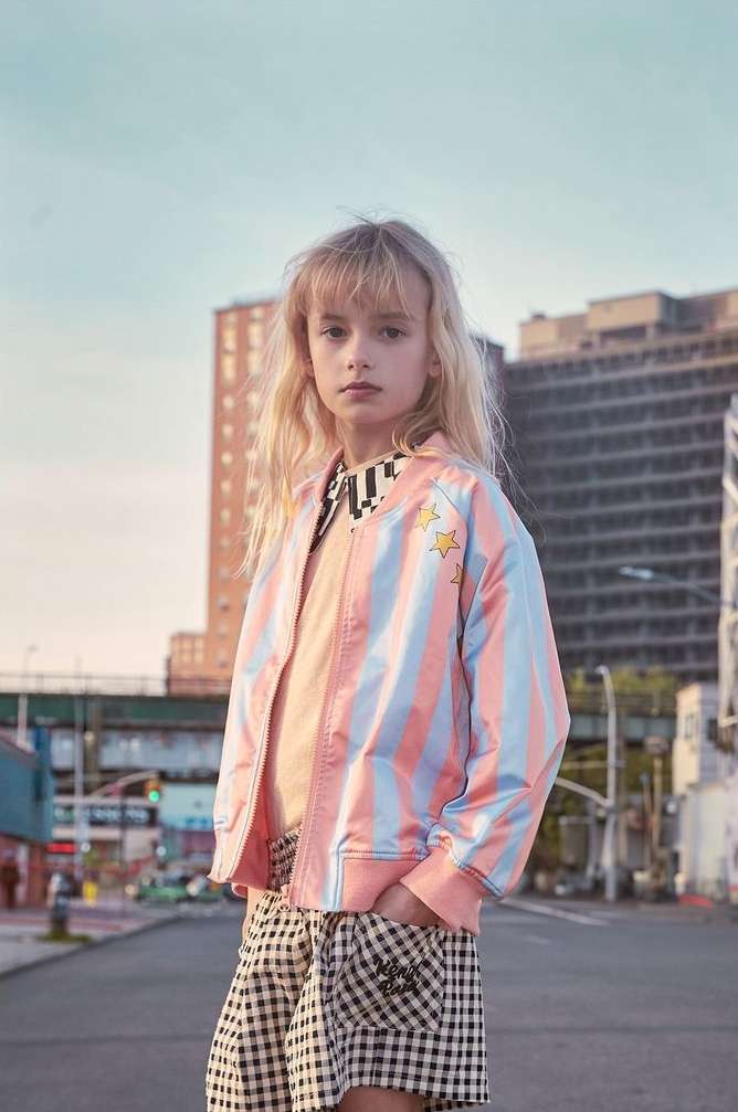 A young girl with blonde hair poses in a pink and blue striped jacket and checkered shorts against an urban backdrop with buildings.