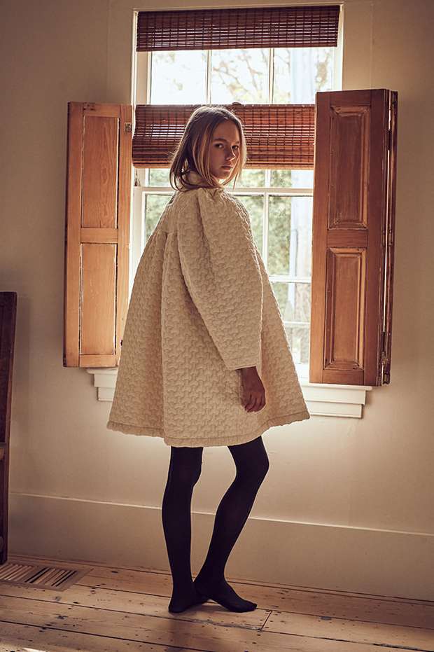 A young woman with blonde hair wearing a cream textured coat and black tights poses in front of a window with open shutters.