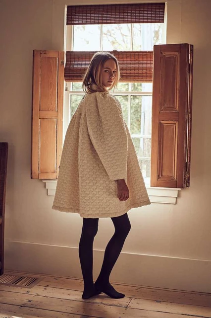 A young woman with blonde hair wearing a cream textured coat and black tights poses in front of a window with open shutters.