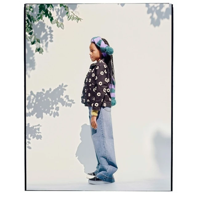A young girl stands in profile, wearing a floral jacket, wide-leg jeans, and a knitted headband with pom-poms, against a white background.