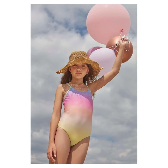 A young girl wearing a straw hat and a pastel-colored swimsuit holds balloons against a partly cloudy sky, creating a summery vibe.