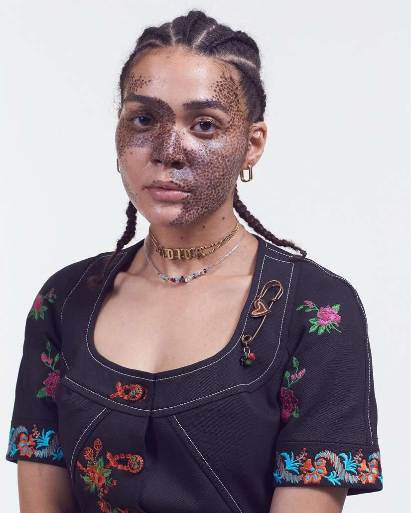Woman with braided hair poses indoors in a black dress with floral embroidery, accessorized with a gold necklace and safety pin.