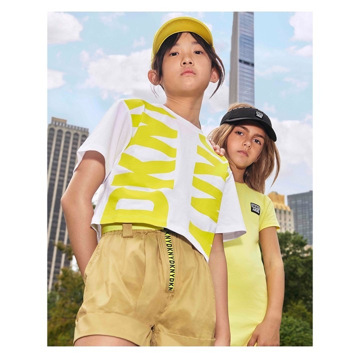 Two children model DKNY clothing in front of a cityscape backdrop, one wears a white and yellow tee, the other in a yellow dress.