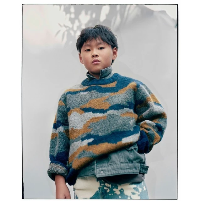 Young child poses indoors wearing a blue, grey, and tan patterned sweater with a denim jacket and tie-dye jeans against a white wall.