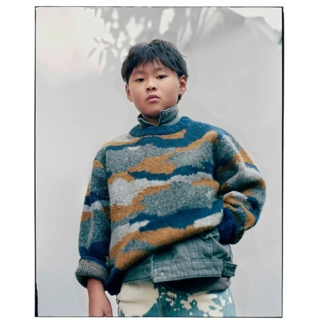 Young child poses indoors wearing a blue, grey, and tan patterned sweater with a denim jacket and tie-dye jeans against a white wall.