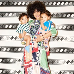 A smiling woman with curly hair holds two toddlers against a patterned black and white Greek key wallpaper background.
