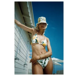 Woman wearing a lemon-patterned bikini and matching bucket hat posing outdoors against a white building wall with a clear blue sky