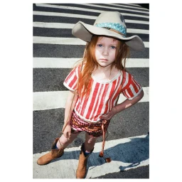 A young red-haired child wearing a cowboy hat, striped t-shirt, patterned shorts, and brown boots standing on a street crosswalk.