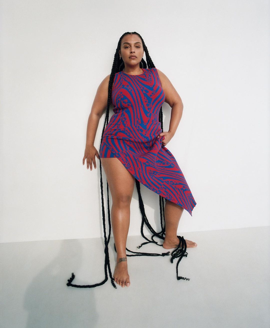 Woman poses barefoot in a patterned dress against a plain white background. Long black braided hair extends down her body and onto the floor.