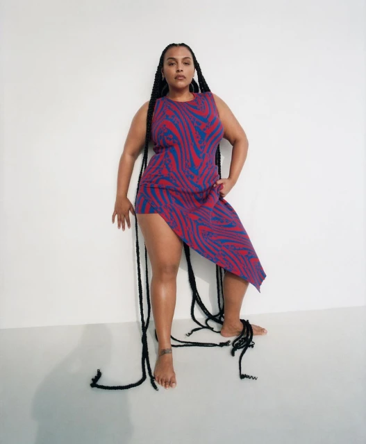 Woman poses barefoot in a patterned dress against a plain white background. Long black braided hair extends down her body and onto the floor.