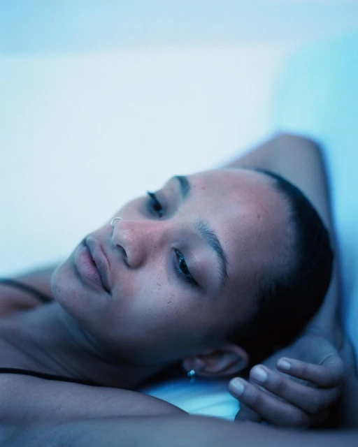 Close-up of a dark-skinned woman lying down with her eyes closed, wearing a black top and silver earrings, against a light blue background.